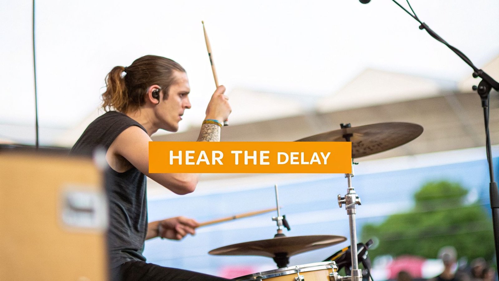 A male drummer with a ponytail and in-ear monitors playing a drum set on stage, with the text 'HEAR THE DELAY'.